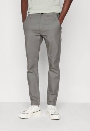 Mężczyzna ubrany w szare, dopasowane spodnie oraz białe, wysokie trampki, stojący z rękami w kieszeniach na jednolitym tle.