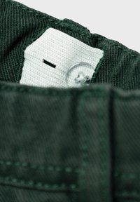 Tessuto verde scuro della cintura dei pantaloni con un'etichetta bianca che presenta un bottone e dettagli di cucitura. Morbida texture con filo visibile.