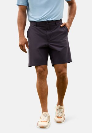 Hombre con pantalones cortos negros, camisa azul claro y zapatillas beige, con una mano en el bolsillo, caminando sobre un fondo claro y liso.