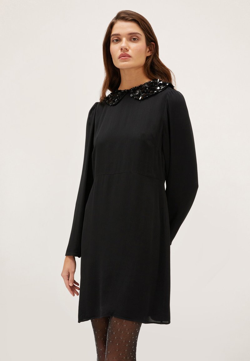 Vestido negro de manga larga con un cuello de lentejuelas, que presenta una silueta recta y una longitud hasta la rodilla, complementado con medias con estampado de puntos.