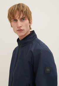Chaqueta azul marina con cuello alto, cierre de cremallera y un parche negro texturizado en la manga. Tejido suave con un brillo sutil.