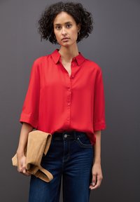 Camicia rossa a maniche corte con bottoni, realizzata in tessuto liscio. Abbinata a jeans blu scuro, con in mano una giacca di suede color beige. Sfondo grigio.