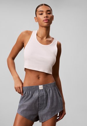 Junge Frau mit zurückgekämmten Haaren, die ein weißes, bauchfreies Tanktop und graue Shorts mit elastischem Bund trägt, steht vor einem grauen Hintergrund.