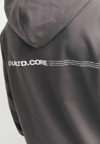 Donkergrijze hoodie met een gladde textuur. Voorzien van witte bedrukte tekst op de achterkant, die het merk en de ontwerp inspiratie beschrijft.