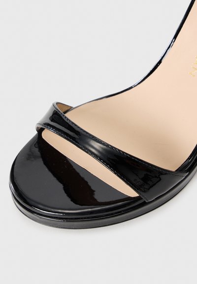 Sandale ouverte en cuir verni noir avec une seule sangle étroite sur les orteils et une semelle intérieure beige sur un fond clair.