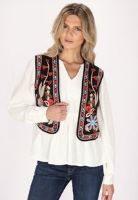 Schwarze bestickte Weste mit floralen Mustern in Rot, Blau und Grün. Getragen über einem weißen langärmeligen Oberteil mit V-Ausschnitt.