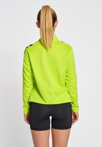 Heldergroen, langemouwen shirt met een kraag en zwarte accenten, gecombineerd met zwarte shorts. Gladde textuur, aansluitend ontwerp en sportieve stijl.