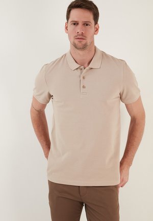 Beige pólóing texturált anyagból, rövid ujjal, három gombos gallérral és egyenes alsó résszel. Barna nadrággal párosítva.