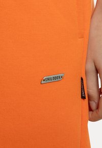 Orange bomuldsstof med en tekstureret overflade. Har et lille sølvtag mærket "SMILODOX" og et sort label på sidesømmen.