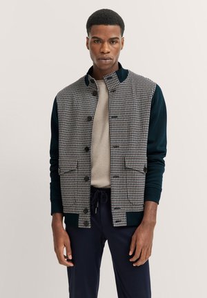Hahnentrittmuster-Jacke mit dunkelgrünen Ärmeln und Kragen. Verfügt über eine Knopfleiste und Fronttaschen. Wird über einem strukturierten beigen Pullover getragen.