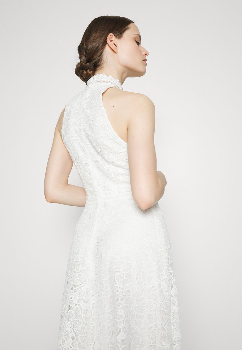 Vestido blanco de encaje con un cuello alto y espalda descubierta. Patrón floral en toda la prenda, con un corsé ajustado y una falda fluida. Textura suave.