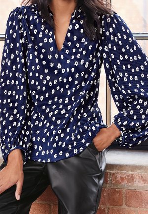 Vrouw draagt een marineblauwe blouse met lange mouwen met witte bloemenprint en zwarte leren broek, zittend met één hand in de zak.