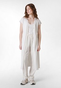 Langer weißer Leinenmantel über einem weißen Tanktop, kombiniert mit hellgestreiften Leinenhosen und weißen Sportschuhen. Mit Knöpfen und einem lockeren Schnitt.
