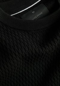 Pull noir en tricot avec un motif texturé, un col côtelé et un subtil accent blanc au-dessus du col. Étiquette de la marque visible.