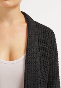 Schwarze Strickjacke mit gerippter Textur und weitem Schalkragen, getragen über einem hellrosa Oberteil, zeigt einen lockeren Schnitt und offene Vorderseite.