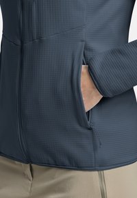 Veste légère bleu foncé avec un motif texturé, fermeture éclair et poches latérales. Les manches ont un poignet ajusté et un tissu doux.