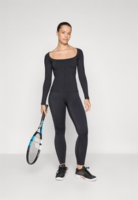 Μαύρο αθλητικό μπλουζάκι με μακριά μανίκια και τετράγωνη λαιμόκοψη, συνδυασμένο με μαύρα εφαρμοστά κολάν. Το μοντέλο κρατά ένα ρακέτα τένις και φοράει μαύρα αθλητικά παπούτσια.