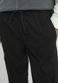Pantalons cargo noirs en tissu léger, avec une taille à cordon, des poches latérales et une texture lisse et mate.