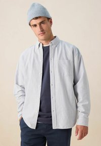 Man in een lichtblauwe gebreide beanie, gestreept overhemd met knopen over een marineblauwe sweatshirt, en donkere broek, staand tegen een effen beige achtergrond.