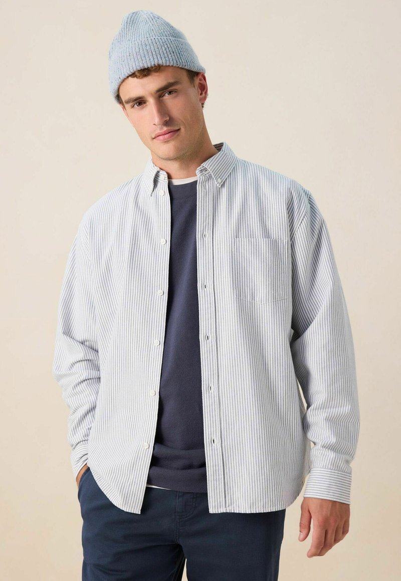 Man in een lichtblauwe gebreide beanie, gestreept overhemd met knopen over een marineblauwe sweatshirt, en donkere broek, staand tegen een effen beige achtergrond.