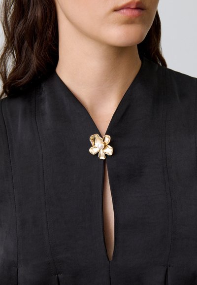 Prenda de tela negra con un profundo escote en V, adornada con un broche metálico en forma de flor y una perla en el centro. Textura suave.