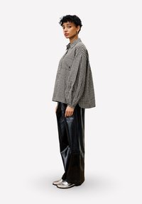 Persoon met kort, krullend haar, gekleed in een oversized zwart-wit geblokt hemd, brede zwarte leren broek en zilveren schoenen, staand in zijaanzicht.