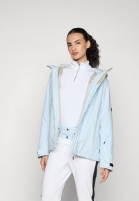 Gaiši zila ūdensnecaurlaidīga jaka ar kapuci, ar rāvējslēdzēju un elastīgām piedurknēm. Iekšpuse ir bēša, ar nelielu jostas detaļu un sānu kabatām.