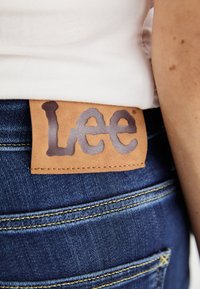 Blå denimjeans med en brun läderetikett på midjebandet, med "Lee"-logotypen i präglad text och kontrasterande stickningar.