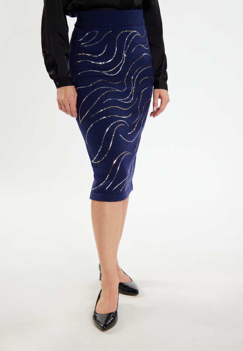 Gonna a matita lavorata a maglia blu navy con motivi ondulati testurizzati, accentuata da dettagli metallici. Abbinata a décolleté nere a punta.