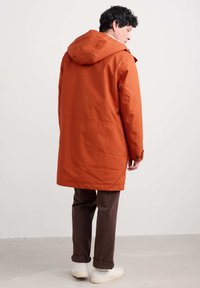 Abrigo impermeable naranja con capucha, de corte recto, con puños abotonados y una abertura trasera, usado sobre pantalones marrones y zapatos blancos.