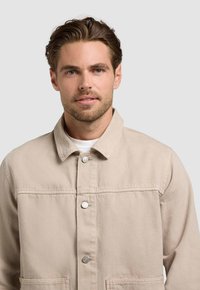 Jeune homme aux cheveux châtain clair et à la barbe, portant une veste beige boutonnée sur une chemise blanche, faisant face à l'avant sur un fond uni.