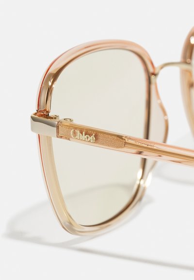 Solglasögon med en transparent persikofärgad ram, grönt tonade linser och en guldaccent på den gångjärnsförsedda armen med varumärket "Chloé."