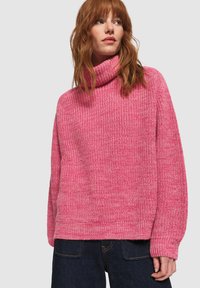 Roze chunky gebreide coltrui met een losse pasvorm, ribbelrand en gestructureerd patroon. Ggedragen met donkere jeans.