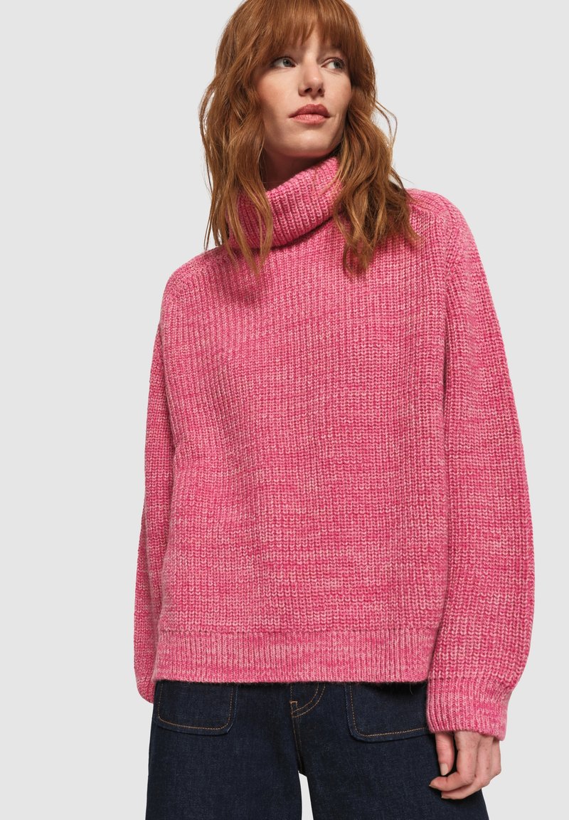 Maglione a collo alto di maglia spessa color rosa, con vestibilità ampia, bordi a costine e motivo texturizzato. Indossato con jeans scuri.