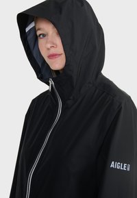 Giacca nera impermeabile con cappuccio, caratterizzata da una zip argento e un logo sulla manica sinistra. Texture liscia con un design minimalista.