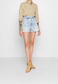 Beige košilová halenka, světle modré džíny s šňůrkou v pase a béžové kotníkové boty. Oblečení má jednoduchý design a jemné textury.