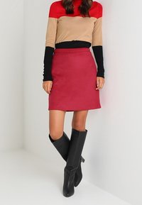 Femme portant un haut à manches longues rouge, beige et noir, une jupe rouge et des bottes noires montant jusqu'aux genoux, se tenant contre un fond blanc.