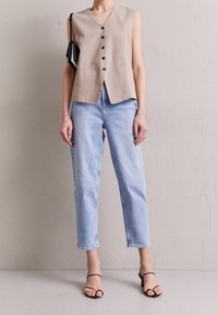 Chaleco sin mangas beige con escote en V y botones negros, combinado con jeans rectos de color azul claro. Sandalias de tacón negras completan el look.