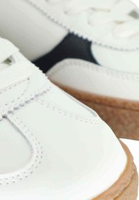 Sneaker bianche in pelle con accenti scuri, dotate di lacci bianchi e suole in gomma robusta color tan, che mostrano dettagli di cucitura intricati.