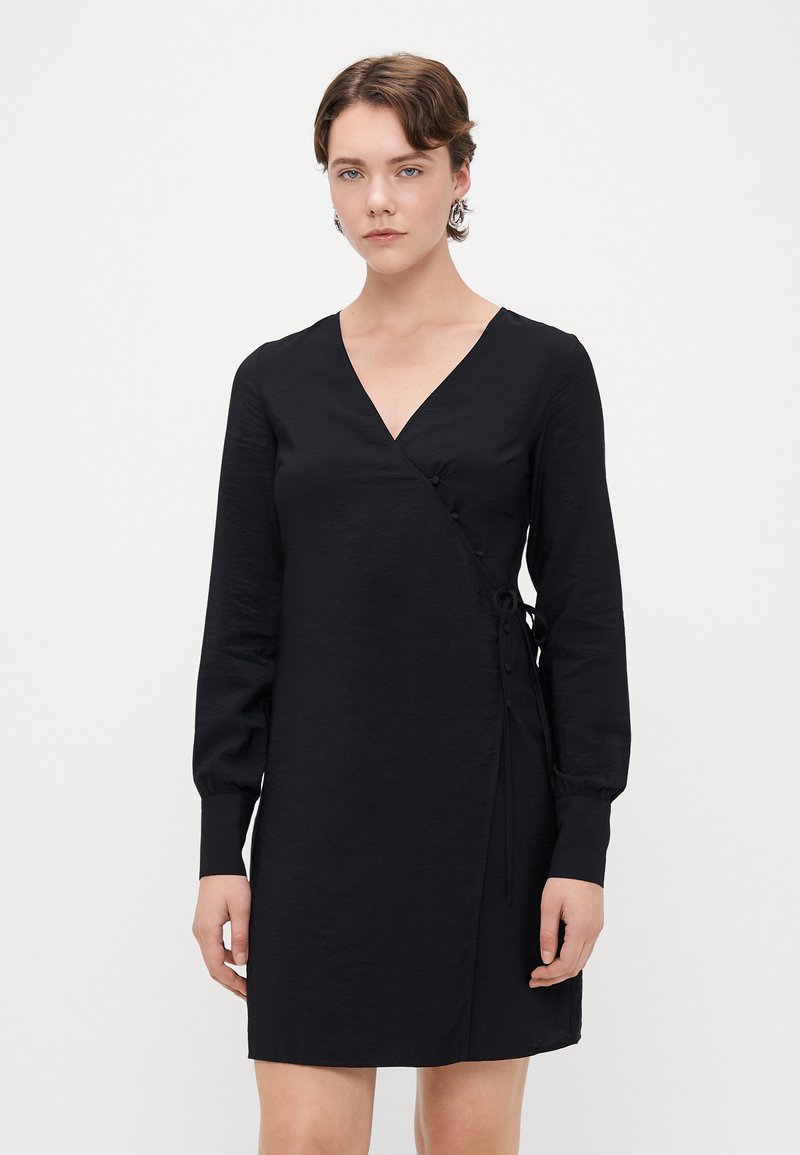 Melnas svārku kleitas ar garām piedurknēm, V-veida izgriezumu, priekšpuses pogu detaļām un sānu saitēm, kas raksturojas ar gludu tekstūru un minimālu dizainu.