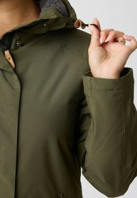 Olijfgroene jas met een gestructureerde capuchonvoering, verstelbare trekkoord, drukknoopsluitingen en schoudernaaddetails.