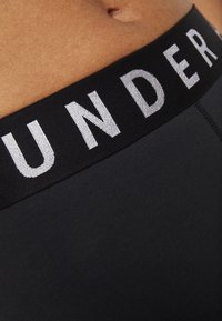 Black fabric underwear with a wide elastic waistband featuring the word "UNDER" in white, subtly raised lettering. Smooth texture, close-up view.