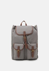 Grå canvasryggsäck med bruna läderdetaljer, två framfickor, en överdel med lock och ett handtag på toppen. Har justerbara remmar och spännband.