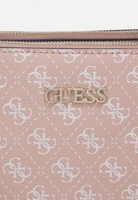 Zbliżenie różowej, teksturowanej torebki z powtarzającym się białym monogramem i złotym logo GUESS obok dwóch złotych zamków błyskawicznych.