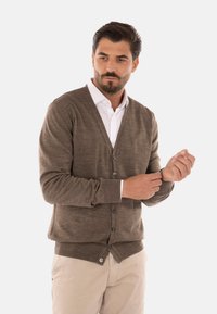 Cardigan in maglia marrone con scollatura a V, chiusura con bottoni e polsini a costine. Indossato sopra una camicia bianca, abbinato a pantaloni chiari.