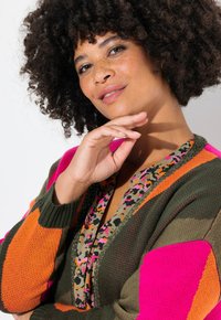 Cardigan en tricot vert, rose et orange, avec un col en V profond et une doublure à motifs animaliers colorés. Manches texturées avec de grands blocs de couleur.