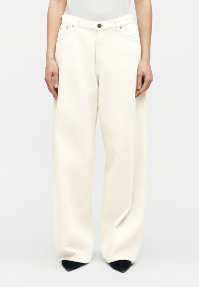 Femme portant un pantalon blanc ample à jambes larges et des chaussures noires pointues, debout devant un fond clair uni.