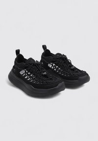 Zapatillas deportivas negras con un diseño de parte superior tejida, suela acolchada y lengüetas de agarre. Presentan una textura suave y acentos metálicos mínimos.