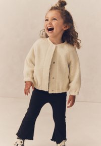 Kremfarvet faux shearling-jakke med knappelukning, kombineret med sorte, udsvingede bukser og hvide sko, med en tekstureret overflade og afrundede manchetter.