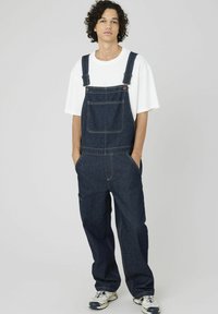 Salopette en jean bleu foncé avec bretelles réglables, poche avant et poches latérales. Associée à un t-shirt blanc et des chaussures de sport.